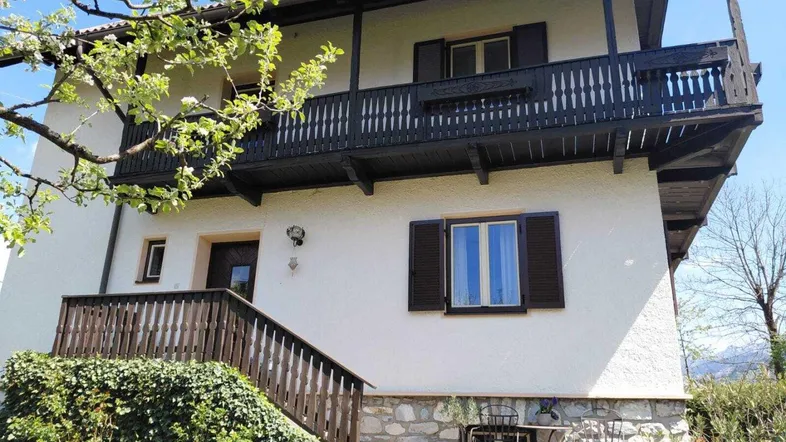 Seitenansicht des Hauses mit Holzbalkon, Steinfundament und gepflegtem Gartenbereich unter blauem Himmel.