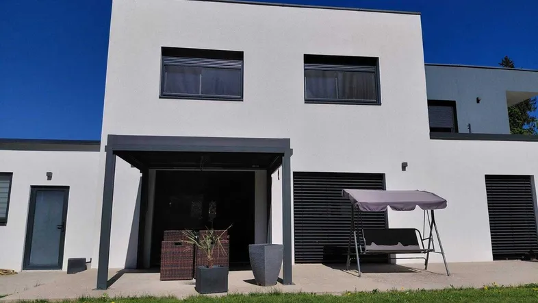 Moderne Hausfassade mit überdachter Terrasse, Rasenfläche und Gartenmöbeln.