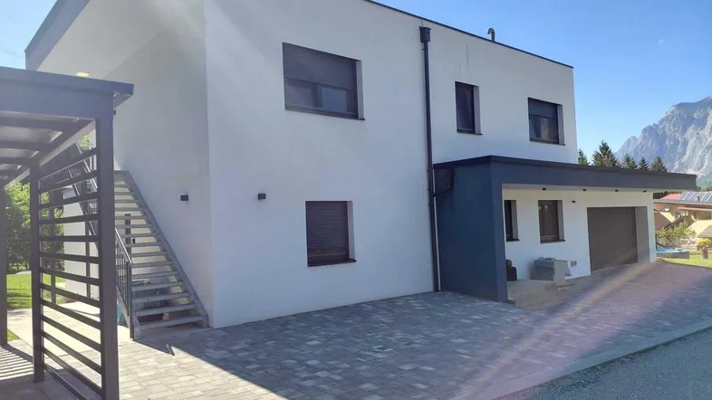 Modernes Einfamilienhaus mit weißer Fassade, Carport und gepflasterter Auffahrt vor Bergkulisse.
