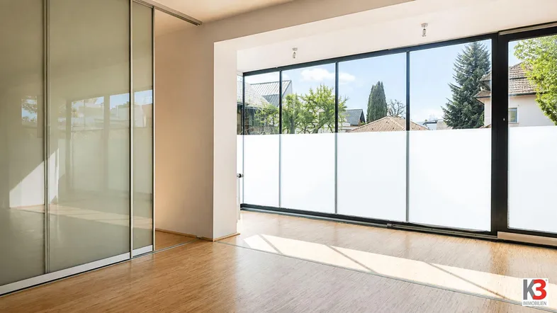 Heller Innenraum mit großen Fenstern, Holzboden und einer modernen Glasschiebetür, die viel Tageslicht hereinlässt.