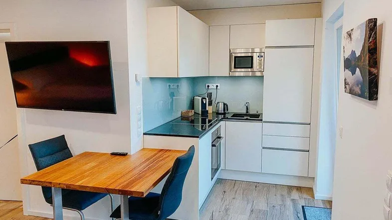 Moderne Küchenzeile mit weißen Schränken, schwarzer Arbeitsplatte und Essbereich mit Fernseher.