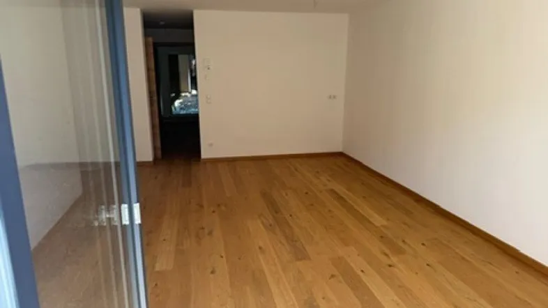 Helles, leeres Zimmer mit Holzboden und Zugang zu einem Balkon oder einer Terrasse.