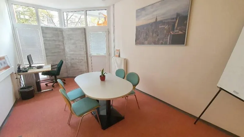 Heller Büroraum mit Teppichboden, Besprechungstisch und großen Fenstern, die viel Tageslicht hereinlassen.