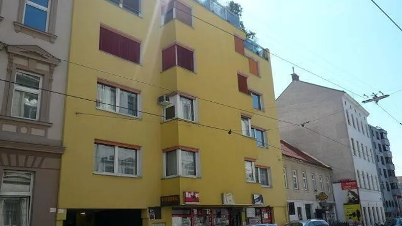 Gelbes Mehrfamilienhaus mit roten Fensterläden und Geschäften im Erdgeschoss an einer belebten Straße.