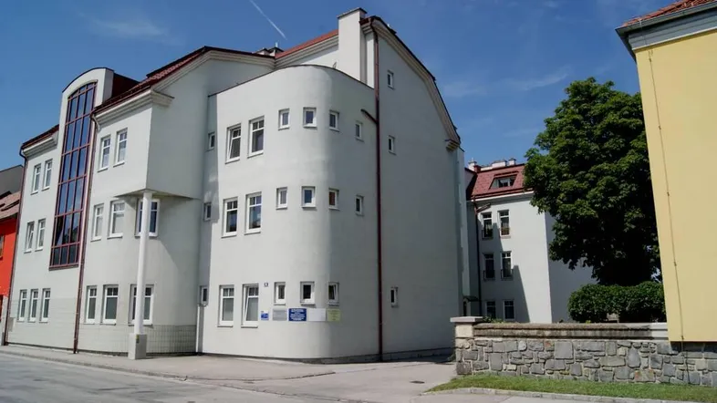 Modernes Gebäude mit heller Fassade, vielen Fenstern und rotem Ziegeldach an einer Straßenecke.
