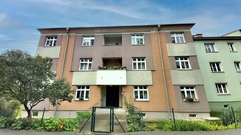 Mehrfamilienhaus mit mehreren Etagen und Balkonen, umgeben von Grünflächen unter blauem Himmel.