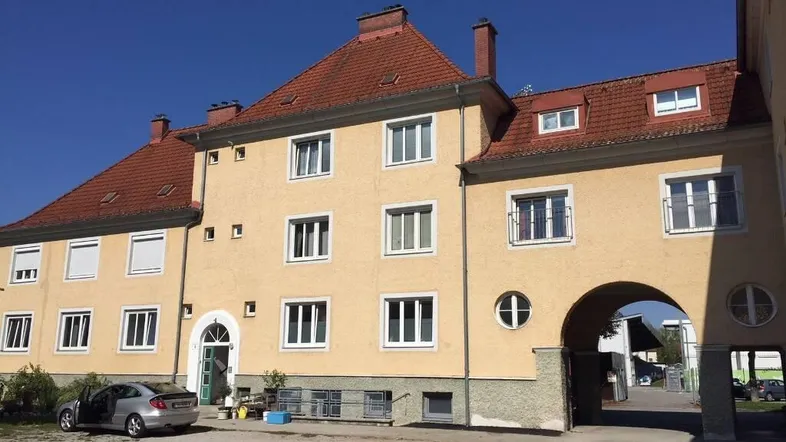 Gelbes Mehrfamilienhaus mit rotem Ziegeldach, weißen Fenstern und einem Torbogen, der zu einem Innenhof führt.