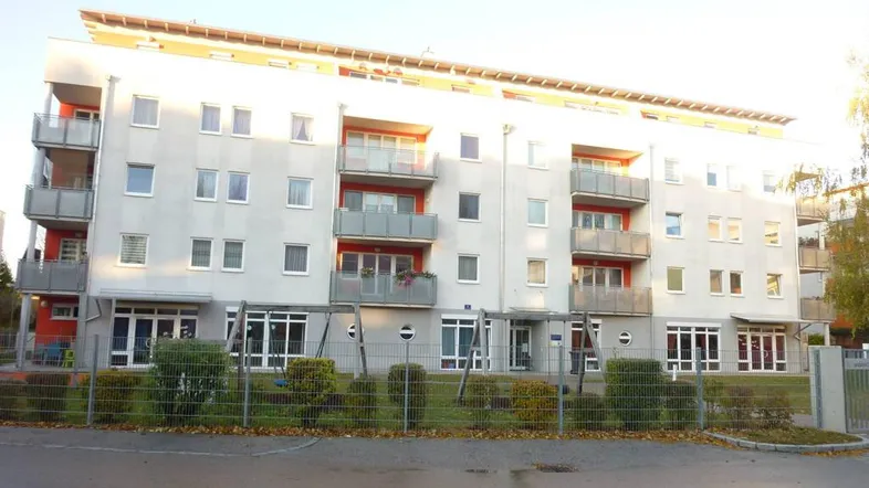 Modernes Mehrfamilienhaus mit hellgrauer Fassade, roten Akzenten und Balkonen sowie einem Spielplatz im Vorgarten.