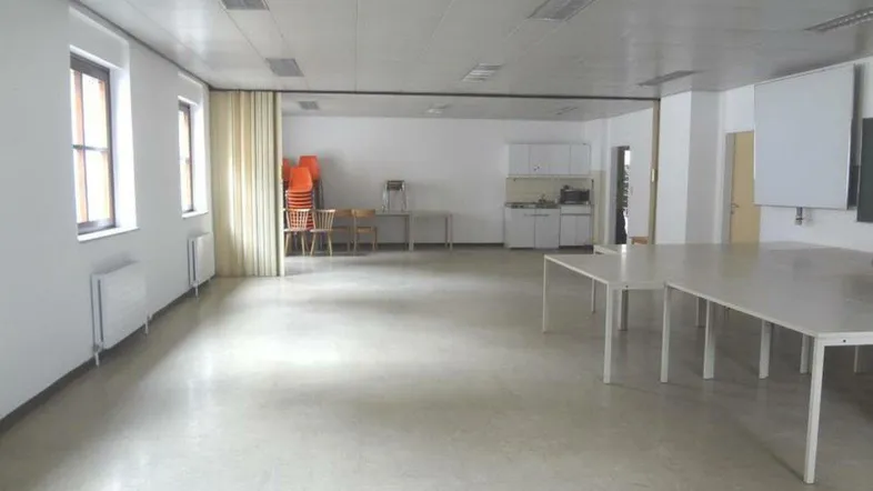 Großer Mehrzweckraum mit mehreren Tischen, einer kleinen Küchenzeile und Fenstern, ideal für Besprechungen.