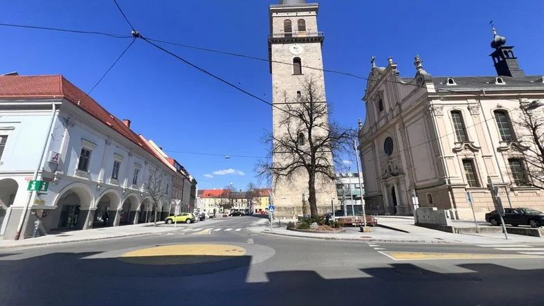 Eine belebte Straße in einer historischen Stadt mit einem markanten Kirchturm und traditionellen Gebäuden unter blauem Himmel.