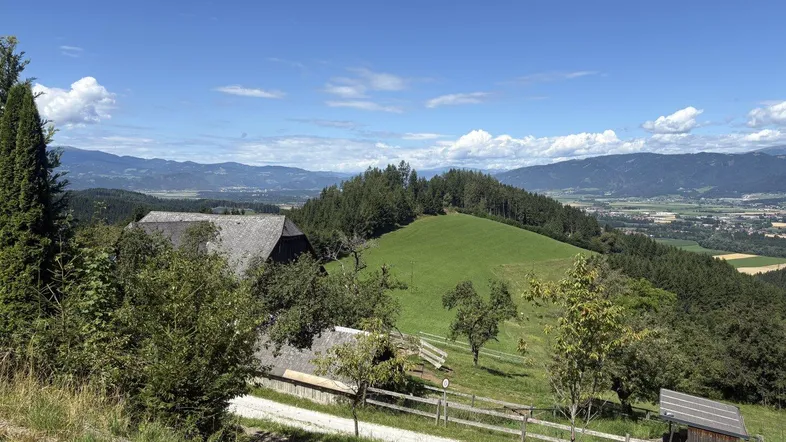 Ländliche Landschaft mit einem Haus, grünen Hügeln und Bergen im Hintergrund unter blauem Himmel.