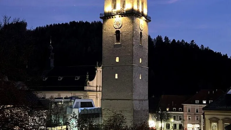 Nachtansicht eines beleuchteten Kirchturms und des Stadtplatzes mit umliegenden Gebäuden und Bäumen.