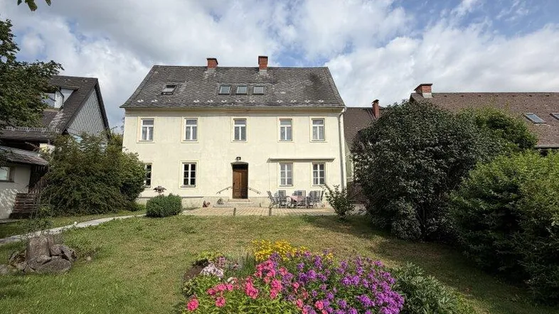 Historisches Gebäude mit gepflegtem Garten, Blumenbeeten und Sitzbereich im Freien unter blauem Himmel.