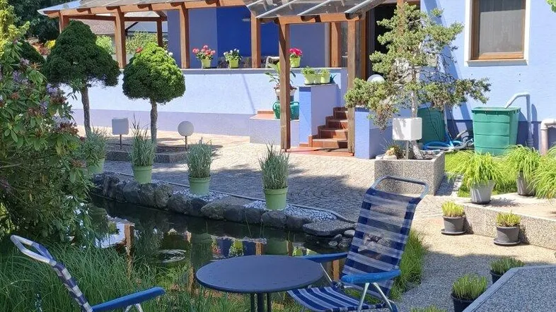 Blauer Hausgarten mit Teich, Sitzgelegenheiten und überdachtem Terrassenbereich unter blauem Himmel.