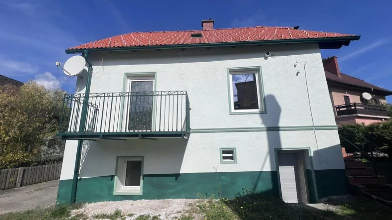 Weißes Haus mit grünem Sockel, rotem Ziegeldach und kleinem Balkon unter blauem Himmel.
