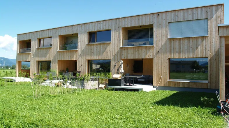 Moderne Holzfassade mit Terrassenbereich und Gartenmöbeln, umgeben von grüner Wiese.