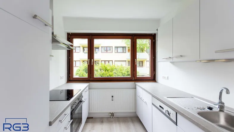 Helle Küche mit weißen Schränken, grauer Arbeitsplatte und Blick ins Grüne durch das Fenster.