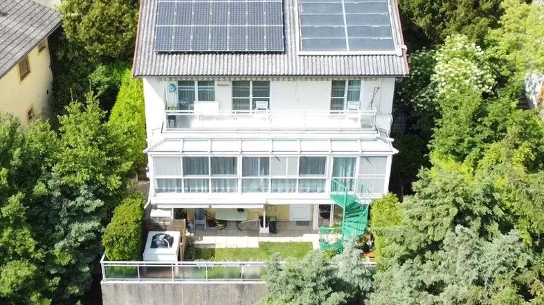 Luftaufnahme eines Hauses mit Solaranlagen auf dem Dach und einem verglasten Wintergarten, umgeben von viel Grün.