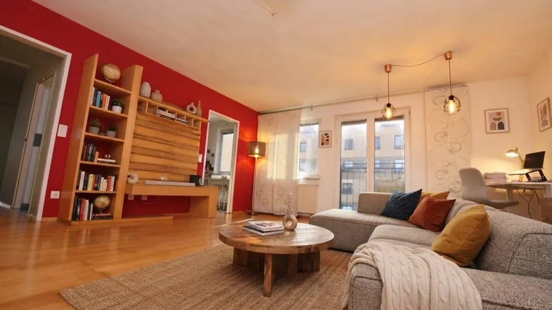 Helles Wohnzimmer mit roter Akzentwand, Parkettboden und Zugang zum Balkon.