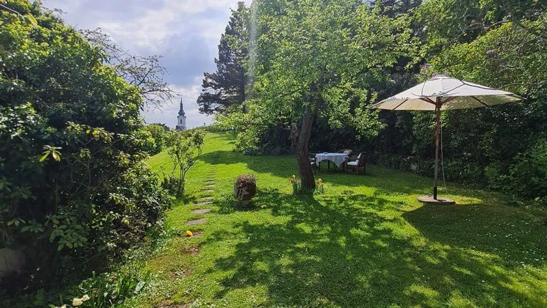 Großer, gepflegter Garten mit Rasenfläche, Sitzgelegenheit und Blick auf eine Kirchturmspitze.