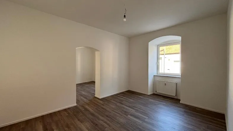 Helles Zimmer mit Laminatboden und einem Bogenfenster, das zu einem weiteren Raum führt.
