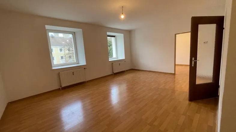 Helles Zimmer mit Parkettboden, weißen Wänden und zwei Fenstern, bereit zur individuellen Gestaltung.