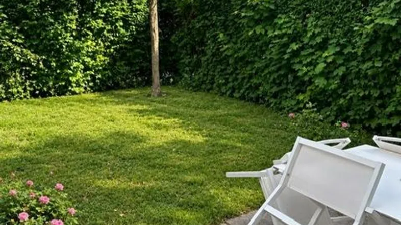 Gepflegter Gartenbereich mit Rasenfläche, hoher Hecke und einer kleinen Terrasse mit Gartenmöbeln.