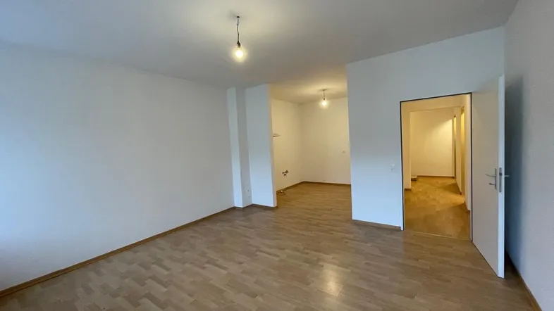 Geräumiges, leeres Wohnzimmer mit hellem Laminatboden und weiß gestrichenen Wänden.