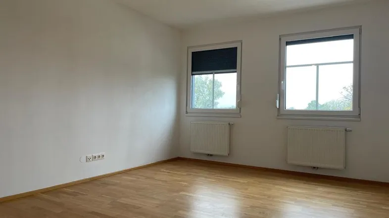 Helles, leeres Zimmer mit Parkettboden, zwei Fenstern und Heizkörpern, bereit zur Einrichtung.