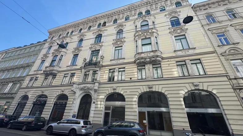 Historisches Gebäude mit reich verzierter Fassade, großen Fenstern und Geschäften im Erdgeschoss an einer Stadtstraße.