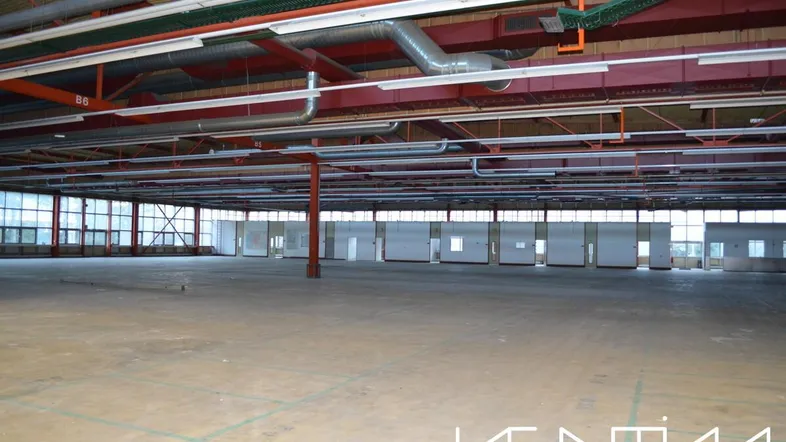 Große, leere Lagerhalle mit hoher Decke, sichtbaren Lüftungsrohren und vielen Fenstern entlang der Wände.