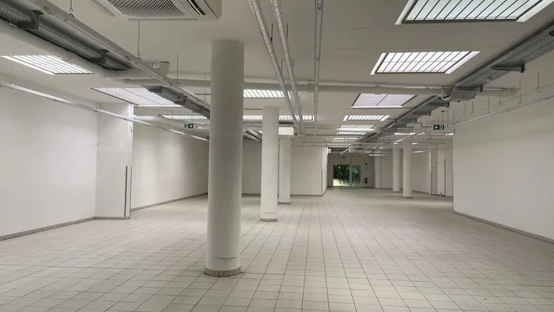 Großer, leerer Gewerberaum mit Fliesenboden, Säulen und sichtbaren Lüftungsrohren an der Decke.