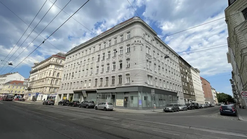 Historisches Eckgebäude an einer belebten Straße mit Geschäften im Erdgeschoss und vielen Fenstern.
