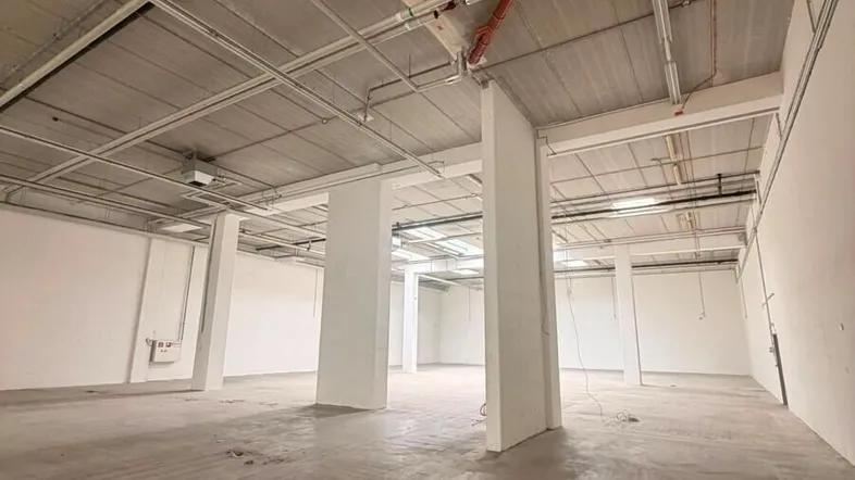Großer, leerer Industrieraum mit Betonboden, sichtbaren Leitungen an der Decke und weißen Wänden.