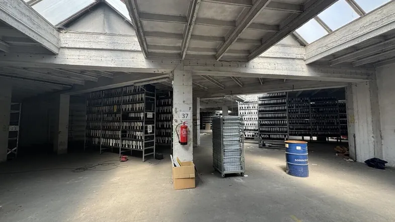 Großer, offener Lagerraum mit Betonstützen, Oberlichtern und Regalen voller Akten oder Flaschen.
