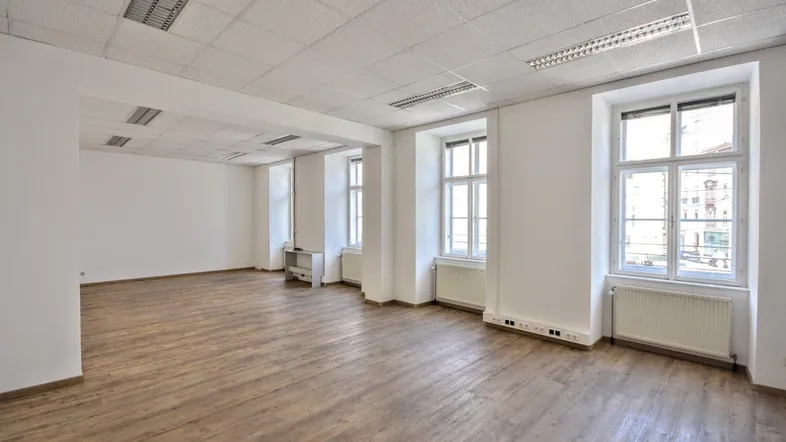 Geräumiger Büroraum mit hellem Laminatboden, weißen Wänden und großen Fenstern, die viel Tageslicht hereinlassen.