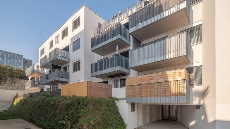Modernes Mehrfamilienhaus mit mehreren Balkonen und begrüntem Hang unter blauem Himmel.