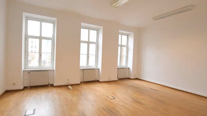 Heller, leerer Raum mit drei großen Fenstern, Holzfußboden und Heizkörpern unter den Fenstern.