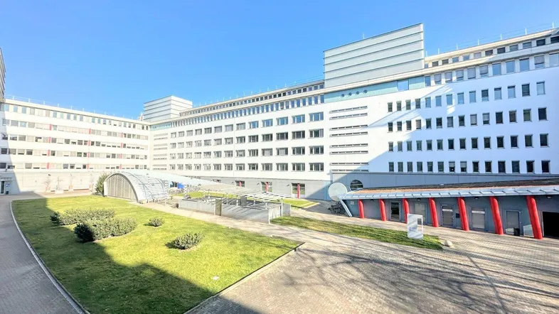 Modernes Gebäude mit weißer Fassade und vielen Fenstern, umgeben von einem gepflasterten Innenhof mit Grünflächen unter blauem Himmel.