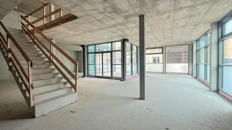 Großer, offener Gewerberaum im Rohbau mit Betondecken, großen Fenstern und einer Treppe.
