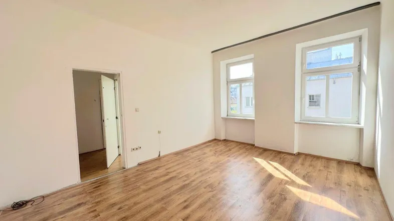 Helles, leeres Zimmer mit Laminatboden und zwei Fenstern, bereit zur individuellen Gestaltung.