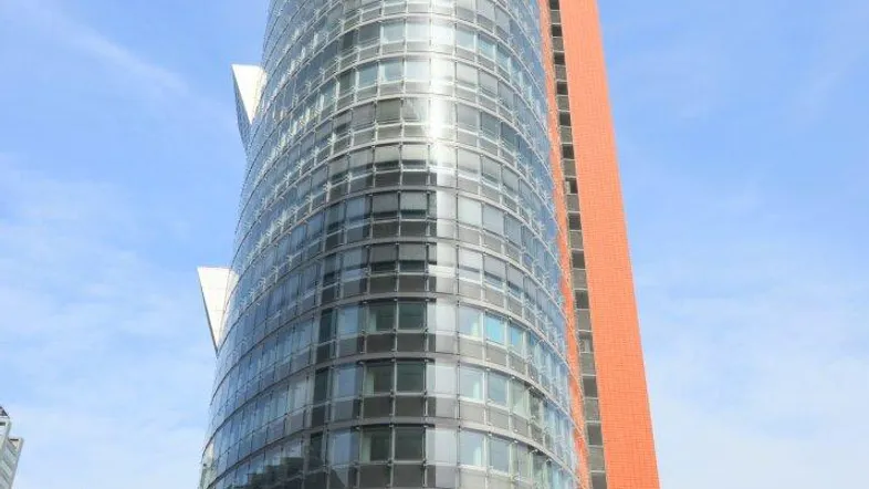 Modernes Hochhaus mit Glasfassade und angrenzendem orangefarbenem Gebäudeteil unter blauem Himmel.