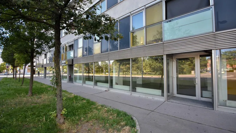 Moderne Gebäudefassade mit großen Glasfenstern im Erdgeschoss und Bäumen entlang des Bürgersteigs.
