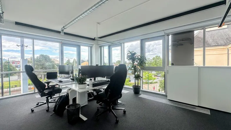 Modernes Büro mit großen Fenstern, Blick ins Grüne und sichtbaren Lüftungsrohren an der Decke.