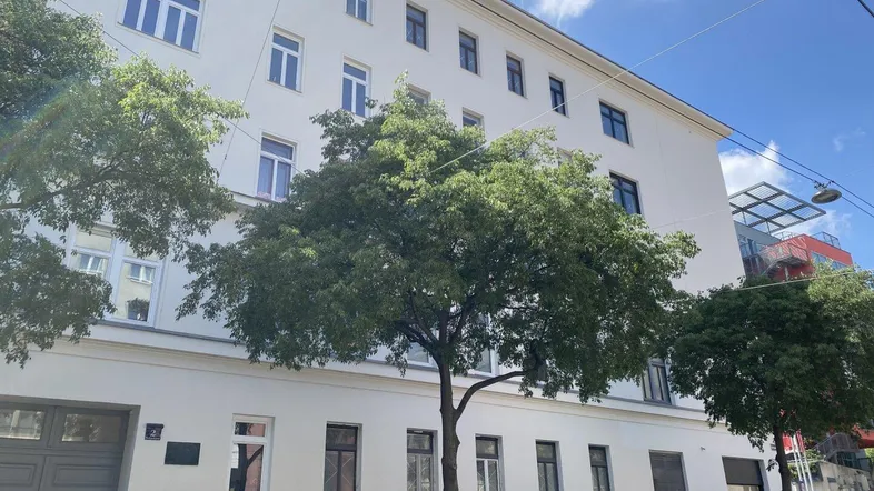 Weißes Mehrfamilienhaus mit vielen Fenstern und Bäumen davor unter blauem Himmel.