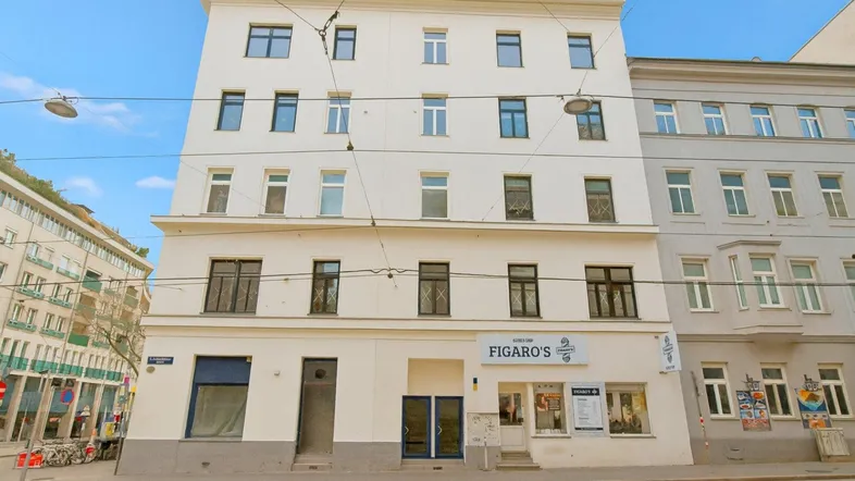 Ansicht eines gepflegten Mehrfamilienhauses mit weißer Fassade und Geschäftsräumen im Erdgeschoss an einer Stadtstraße.