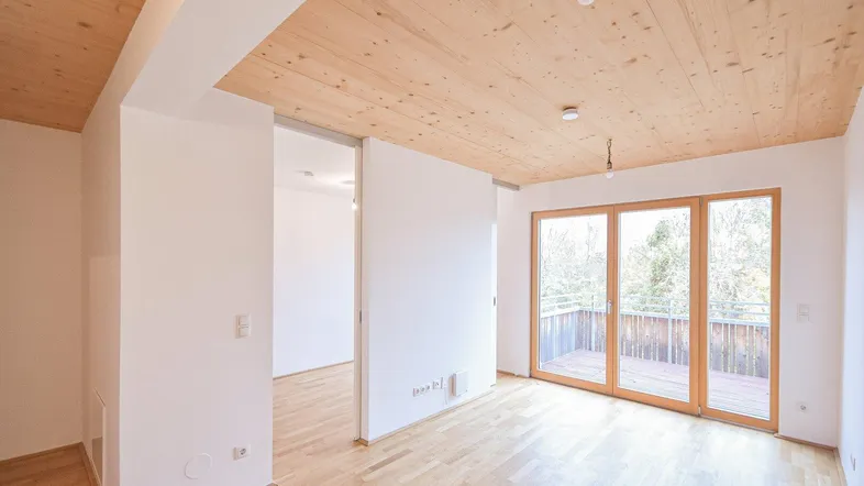 Helles Zimmer mit Holzboden und Holzdecke, großen Fenstern und Zugang zu einem Balkon.