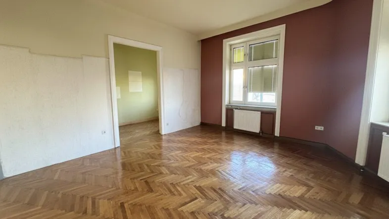 Geräumiges Zimmer mit Fischgrätparkett und hohen Fenstern, bereit zur Renovierung.