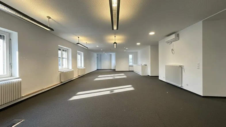 Geräumiger, leerer Büroraum mit Teppichboden, vielen Fenstern und moderner Beleuchtung.