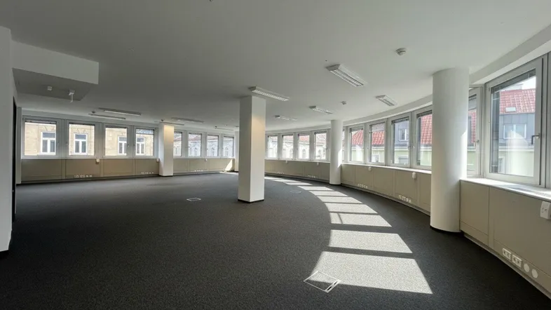 Geräumiger, leerer Büroraum mit Teppichboden und vielen Fenstern, die viel Tageslicht hereinlassen.